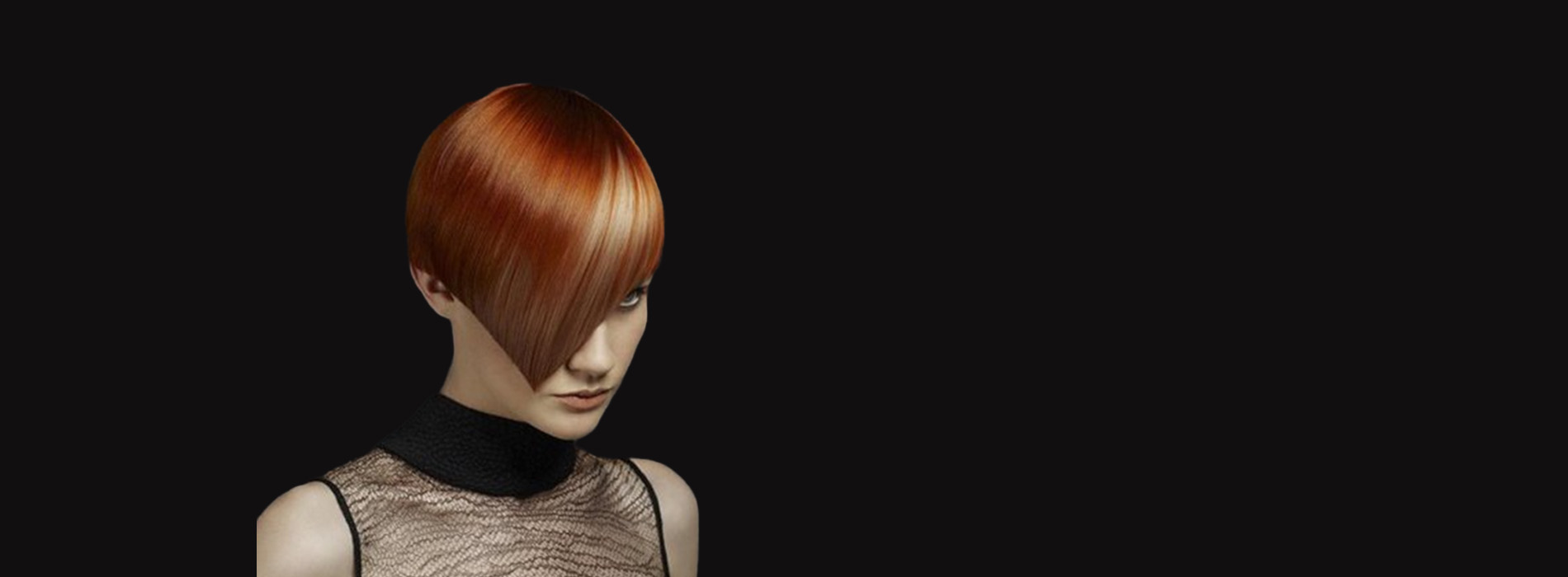 The image shows a person with short hair, wearing a black top and a necklace. They are standing against a dark background.